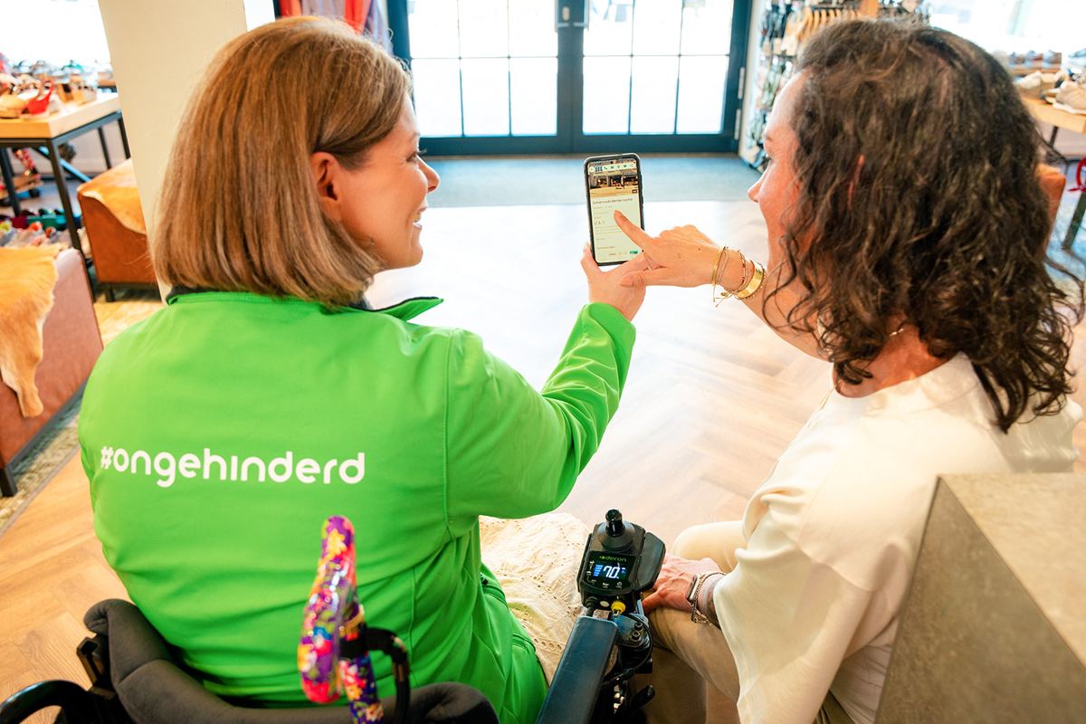 Een ambassadeur van Ongehinderd in een rolstoel laat een ondernemer iets zien op haar telefoon, in een winkel.