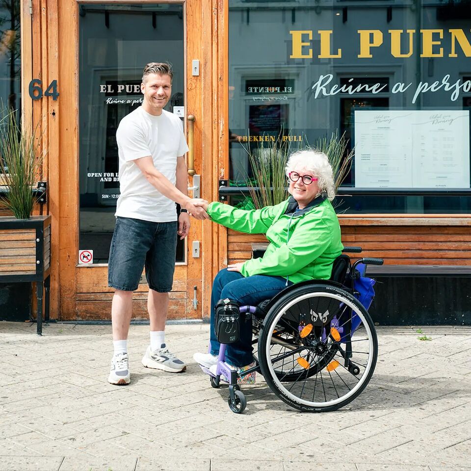 Een staande man schudt de hand van een vrouw in een rolstoel, voor de ingang van een restaurant.